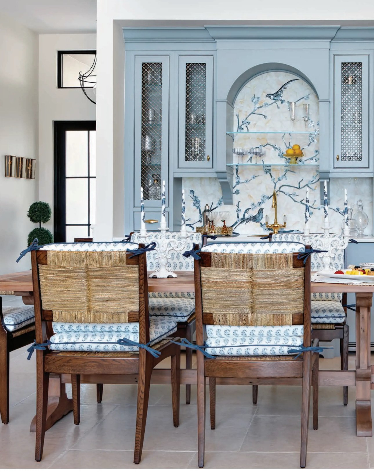 Dining room with rush-seat chairs in front of blue cabinetry and the arched bird and blossom mosaic niche