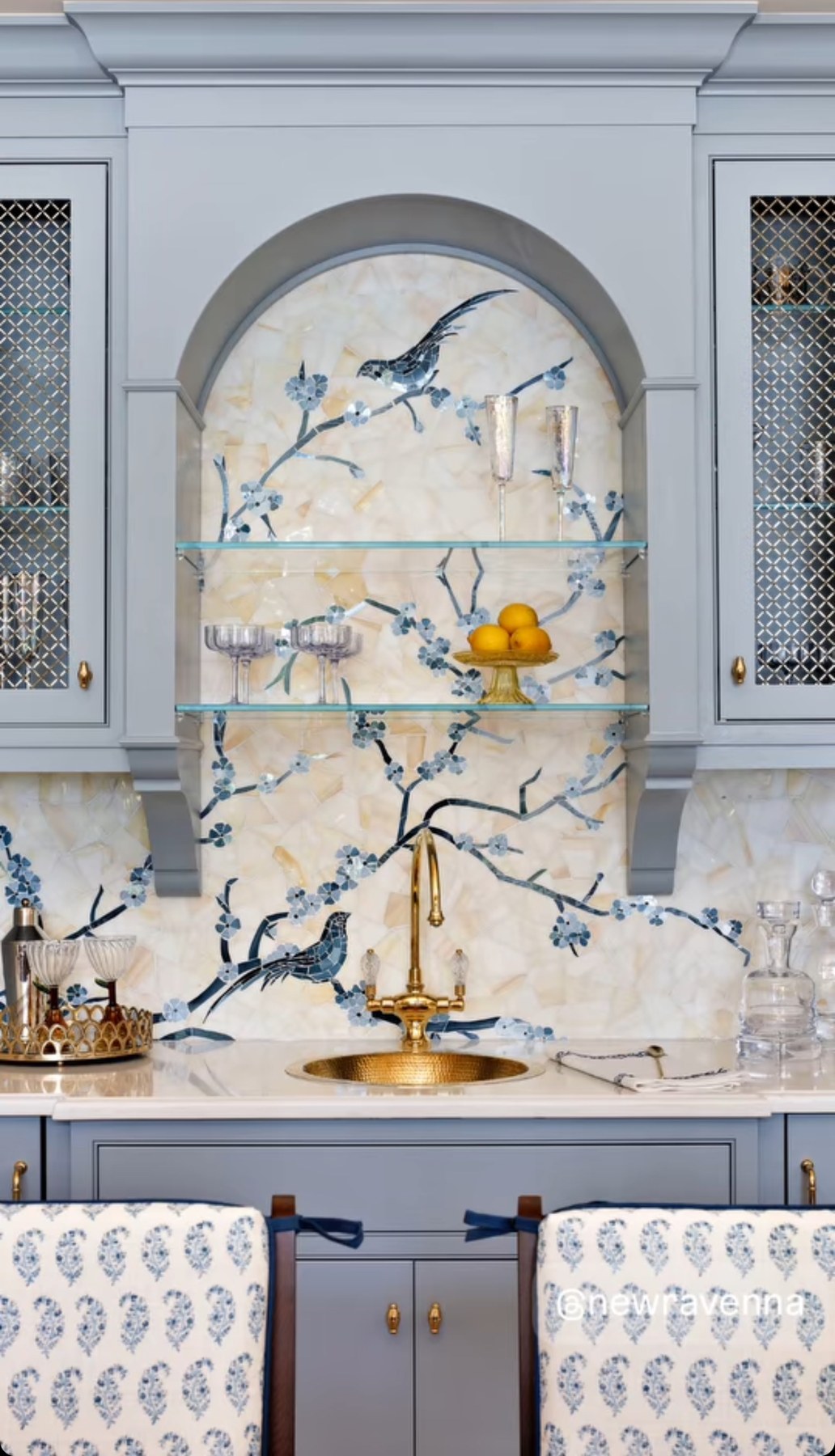 Arched niche in blue-grey cabinetry lined with a mosaic of blue blossoming branches and two birds on a pale stone ground, above a brass bar sink